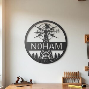 Peut inclure: Une décoration murale en métal noir avec un design circulaire. Le design comprend le nom "NOHAM", une éolienne stylisée et le texte "EST.2026". L'œuvre comprend une silhouette de forêt en bas. Accrochée sur un mur blanc, idéale pour la décoration intérieure.