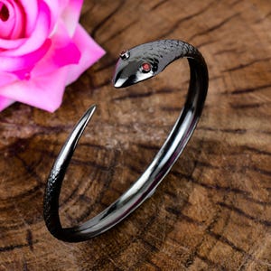 Puede incluir: Un brazalete de metal negro con forma de serpiente enroscada, con escamas texturizadas a lo largo del cuerpo y dos pequeñas piedras redondas de color rojo incrustadas como ojos en la cabeza. El brazalete tiene un diseño abierto y ajustable.