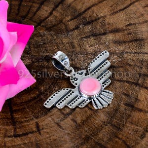 May include: A sterling silver pendant shaped like a bird with outstretched wings, featuring a round, smooth pink stone set in the centre. The silver surface is decorated with small engraved dots and lines, and the bail has sun-like symbols.