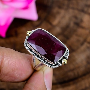 May include: A rectangular faceted deep red ruby gemstone set in a silver ring with a twisted rope border. The setting features two small gold-toned spherical accents on opposite sides of the stone, mounted on a polished silver band.