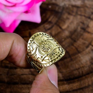 Op de afbeelding: Een goudkleurige metalen ring met een ingewikkeld reliëfontwerp op de brede band. Het oppervlak toont een centraal cirkelvormig motief omringd door geometrische patronen, waaronder zonnestraalachtige lijnen en gekartelde randen aan de boven- en onderkant.