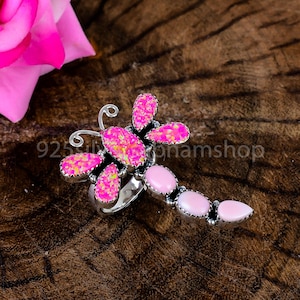 May include: A silver ring shaped like a dragonfly featuring bright pink iridescent wings and a tail composed of three smooth, light pink oval stones. The wings have a textured, sparkling appearance, while the tail stones are solid and opaque.