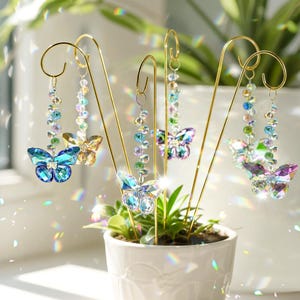 Può includere: Ornamenti decorativi a forma di farfalla in vari colori, tra cui blu, oro e viola. Le farfalle sono realizzate in cristallo sfaccettato e sono appese a supporti metallici dorati. Gli ornamenti sono disposti in un vaso di ceramica bianca con piante verdi.
