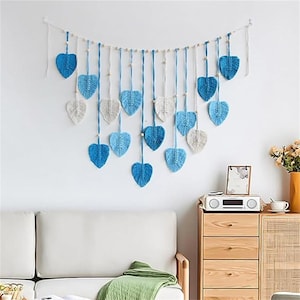 May include: A wall hanging featuring blue and off-white heart-shaped decorations. The hearts are suspended from a white string with wooden beads. The decor is displayed against a white wall, above a white couch.