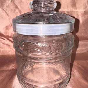 May include: A clear glass jar with a lid. The jar has a decorative, textured pattern around the body and lid. The lid has a white band and a faceted knob. The jar is likely used for storage.