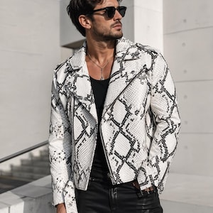 May include: A white and black snakeskin-patterned leather jacket with a zip closure. The jacket has a classic biker style with a notched collar and silver hardware. The model is wearing black sunglasses and black trousers.