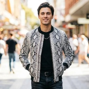May include: A bomber jacket featuring a grey and black snakeskin pattern, a black ribbed collar, black ribbed cuffs, and a front zipper closure. The jacket is worn over a plain black t-shirt, paired with dark denim jeans and a black belt.