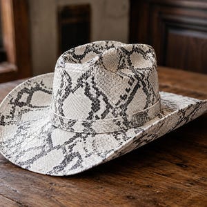 Puede incluir: Un sombrero de vaquero con un patrón de piel de serpiente en blanco y negro en toda la copa y el ala. El sombrero incluye una banda de piel de serpiente a juego alrededor de la base de la copa, acentuada con un emblema metálico plateado circular.