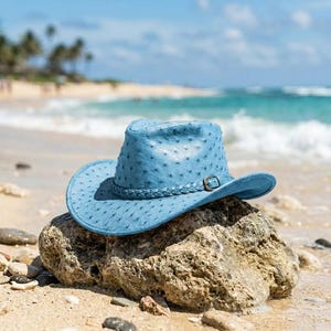 May include: A light blue cowboy hat with a textured, dotted surface pattern. The hat features a braided band around the crown and a small metal buckle detail on the side. The brim is curved upwards on the sides, creating a classic western silhouette.