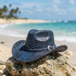 May include: A dark blue cowboy hat featuring a textured snakeskin pattern across the crown and brim. The hat includes a braided band around the base and a metallic buckle detail on the side, showcasing a structured design with a curved brim.