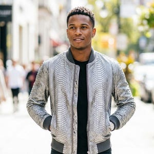 May include: A silver bomber jacket with a textured snakeskin pattern, featuring a dark grey ribbed collar, cuffs, and hem. The jacket is worn over a plain black t-shirt and is designed with a front zip closure and two side pockets.