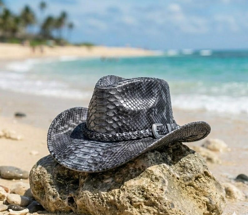 May include: A dark gray cowboy hat with a textured snakeskin pattern. The hat features a braided band around the crown and a metallic buckle detail on the side. The brim is curved upward on the sides, and the crown has a central crease.