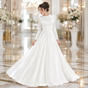 Pode incluir: Um vestido de noiva branco longo com saia ampla e mangas compridas. O vestido tem um corpete justo e um decote redondo. A noiva está em um grande salão com arranjos florais e lustres.