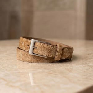 Puede incluir: Un cinturón de piel con pelo en tonos beige y marrón con un acabado de textura natural. El cinturón incluye una hebilla rectangular de metal plateado cepillado y una trabilla a juego, enrollado en forma circular para su exhibición.