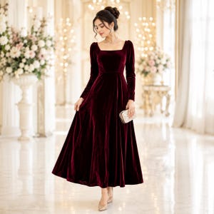 Può includere: Un lungo abito in velluto bordeaux con scollatura quadrata e maniche lunghe. L'abito ha una gonna svasata ed è abbinato a una piccola pochette ornata e tacchi abbinati. L'ambientazione è una stanza elegante e ben illuminata con composizioni floreali.