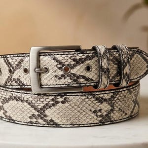 Op de afbeelding: Een opgerolde riem met een slangenleerpatroon in crème, grijze en zwarte tinten. De riem is voorzien van een rechthoekige gesp van geborsteld zilverkleurig metaal, bijpassende metalen oogjes en twee lussen om de riem op zijn plek te houden.