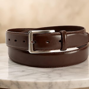 May include: A dark brown leather belt with a smooth, polished finish and a rectangular silver-toned metal buckle. The belt features a single matching leather keeper loop and a series of evenly spaced adjustment holes along the strap length.