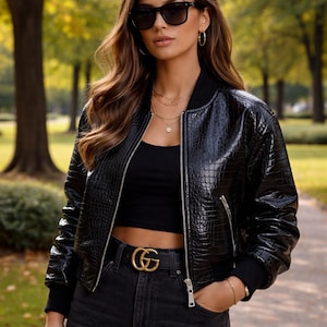 Puede incluir: Chaqueta bomber de cuero negro texturizado con cierre de cremallera. La chaqueta se combina con un top negro, vaqueros oscuros y un cinturón dorado. La persona lleva gafas de sol y joyas doradas.