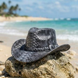 May include: A dark gray cowboy hat with a textured snakeskin pattern. The hat features a braided band around the crown and a metallic buckle detail on the side. The brim is curved upward on the sides, and the crown has a central crease.