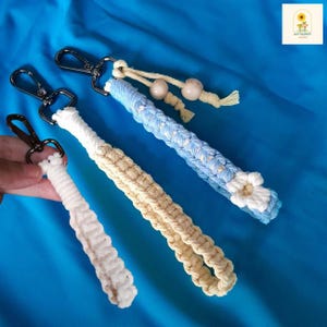 Op de afbeelding: Drie macramé polsband sleutelhangers in wit, beige en lichtblauw. Elke sleutelhanger heeft een geweven ontwerp en een metalen sluiting. De blauwe sleutelhanger heeft een witte bloem en houten kralen. De sleutelhangers worden gepresenteerd op een blauwe stoffen achtergrond.