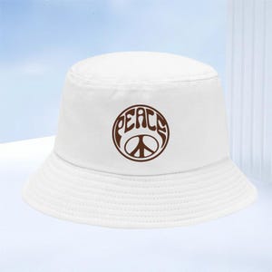 Puede incluir: Un sombrero de pescador blanco con un gráfico circular marrón en la parte delantera. El diseño muestra la palabra PEACE escrita en una fuente estilizada sobre un símbolo de la paz. El sombrero tiene una copa plana y un ala inclinada hacia abajo.