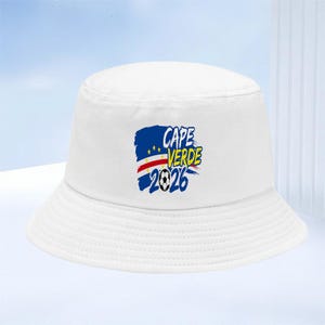 Pode incluir: Um chapéu bucket branco com um design gráfico nas cores da bandeira de Cabo Verde, o texto "CAPE VERDE 2026" e uma ilustração de uma bola de futebol. O design está centrado na parte frontal do chapéu, que possui uma aba larga e inclinada.