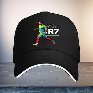 Puede incluir: Una gorra de béisbol negra con un gráfico colorido de un jugador de fútbol en movimiento. El diseño incluye el texto RONALDO y CR7 en letras blancas, con pequeños acentos rojos, amarillos y verdes situados sobre y bajo el gráfico.