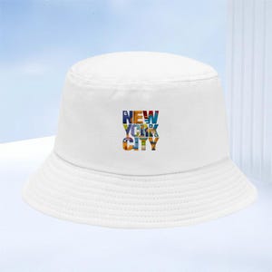 Pode incluir: Um chapéu bucket branco com um design gráfico centralizado com o texto NEW YORK CITY. As letras são preenchidas com uma estampa de mosaico colorida e com vários padrões, contrastando com o tecido branco sólido da copa e da aba.