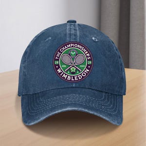 May include: A blue denim baseball cap featuring a circular logo on the front. The logo displays two crossed tennis rackets, a tennis ball, and the text THE CHAMPIONSHIPS WIMBLEDON with the numbers 28 on both sides of the central graphic.