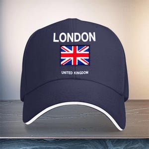 Pode incluir: Um boné de beisebol azul-marinho com a palavra LONDON em letras maiúsculas brancas sobre uma bandeira Union Jack bordada. Abaixo da bandeira, o texto UNITED KINGDOM está impresso em branco. O boné tem um acabamento branco na aba.