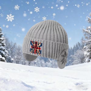 Pode incluir: Um gorro de malha canelada cinza-claro com abas para as orelhas. A parte frontal apresenta um emblema retangular com a bandeira Union Jack ao fundo e o texto PONKS NOT DEAD em letras pretas em negrito.