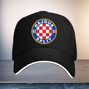 May include: A black baseball cap featuring a circular emblem on the front. The emblem displays a red and white checkered pattern inside a blue ring with the text HAJDUK SPLIT in white letters. The cap has a white trim along the edge of the brim.