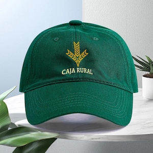 Pode incluir: Um boné de beisebol verde-escuro com um gráfico bordado em amarelo de uma espiga de trigo estilizada acima do texto CAJA RURAL em letras maiúsculas amarelas. O boné tem uma copa estruturada, aba curva e um botão no topo central.