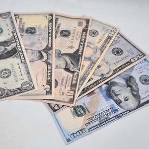 May include: A fan of United States paper currency including a one-dollar bill, a five-dollar bill, a ten-dollar bill, a twenty-dollar bill, a fifty-dollar bill, and a one-hundred-dollar bill, all arranged in a layered, overlapping horizontal display.