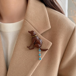 Pode incluir: Um broche de cão castanho com uma coleira dourada e uma corrente prateada pendente com três pequenos cristais azul-claros em forma de coração. O cão está representado numa posição sentada com a cauda estendida para a corrente.