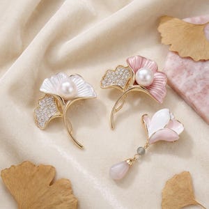 May include: Three elegant brooches in the shape of ginkgo leaves. Two are white and pink with pearl accents and gold stems, and one is a pendant with a pink teardrop. The brooches are set on a cream-colored fabric.