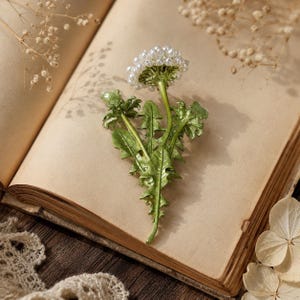 Pode incluir: Um broche de dente-de-leão com um grupo de pequenas contas brancas esféricas representando a flor, fixado a um caule e folhas serrilhadas de metal esmaltado verde. A peça apresenta um acabamento brilhante na folhagem.
