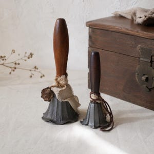 May include: Two hand bells with dark grey fluted metal bodies and smooth wooden handles. Each bell is adorned with a piece of frayed beige fabric tied with twine, and the larger bell features a small, circular, dark brown wax-like seal attached.