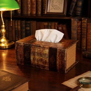 Könnte beinhalten: Eine rechteckige Taschentuchbox im Design eines antiken, ledergebundenen Buches mit abgenutzter brauner Oberfläche. Auf dem Buchrücken steht der Text "PHYTOGRAPHIA CURIOSA" in schwarzen Buchstaben. Weiße Taschentücher ragen aus der Öffnung.