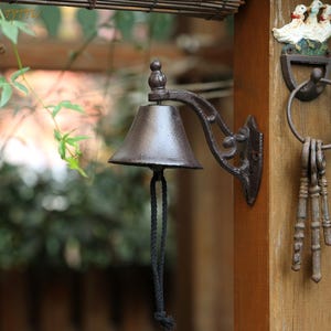 Pode incluir: Um sino de ferro fundido de cor bronze com um puxador de corda preta, montado num poste de madeira. O sino tem um suporte ornamentado e está ao lado de uma placa decorativa de pato e um conjunto de chaves.