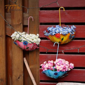 May include: Three colorful, umbrella-shaped hanging planters filled with flowers. The planters are painted in shades of pink, yellow, and blue, and are decorated with floral and butterfly designs. The planters are suspended from hooks and are against a red wooden fence.