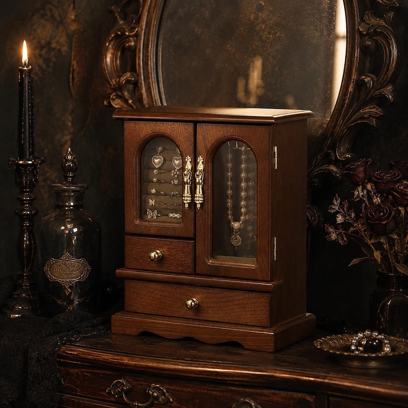 Op de afbeelding: Een donkerbruine houten sieradenkast met twee glazen deuren met goudkleurige handgrepen, een kleine lade en een grote onderste lade. Het interieur toont diverse oorbellen en een ketting die achter de glazen panelen hangen.