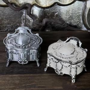 Pode incluir: Duas caixas de joias ornamentadas, de cor prateada, com intrincados desenhos florais e de volutas. Uma é prateada fosca, a outra branca com detalhes prateados. Ambas têm tampas curvas e pés pequenos, sobre uma superfície de madeira escura.