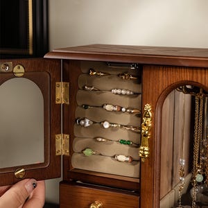 Op de afbeelding: Een houten sieradenkast met een openslaande deur voorzien van een boogvormige spiegel. Het interieur bevat een beige fluwelen ringhouder met diverse ringen, een kleine lade met een gouden knop en een glazen compartiment voor kettingen.