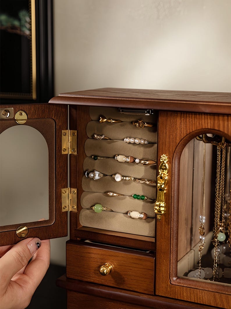 Op de afbeelding: Een houten sieradenkast met een openslaande deur voorzien van een boogvormige spiegel. Het interieur bevat een beige fluwelen ringhouder met diverse ringen, een kleine lade met een gouden knop en een glazen compartiment voor kettingen.