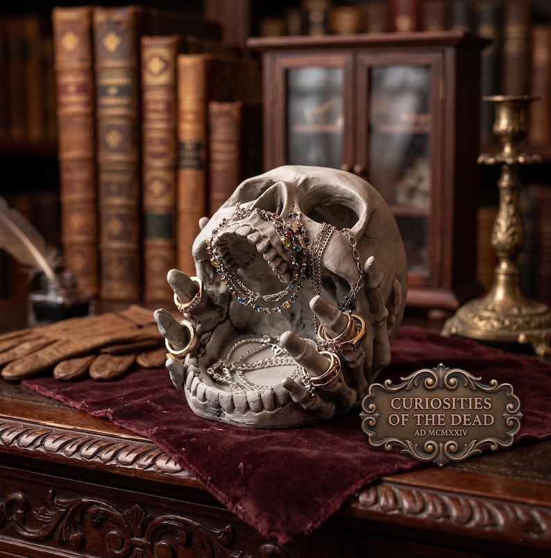 May include: A grey skull-shaped jewellery holder with rings and necklaces. The skull has open jaws and outstretched hands, holding jewellery. A sign reads "Curiosities of the Dead AD MCMXIV". The background includes books and a small cabinet.