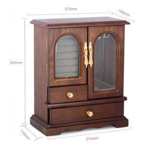 Op de afbeelding: Een donkerbruine houten sieradenkast met twee lades, goudkleurige knoppen en twee glazen deuren. De kast is 215 mm breed, 109 mm diep en 262 mm hoog. Het interieur is voorzien van een grijze fluwelen voering en een rek voor accessoires.