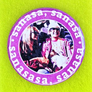 May include: A circular pin-back button featuring a photographic image of two people, surrounded by a purple border with the repeated white text 'sanasa,'. The button is mounted on a lime green display card with 'HAQ JOB media' and 'Niche gifts for radical human beings' printed in black.