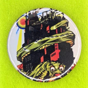 May include: A circular pin-back button featuring a graphic illustration of a green creature wrapped around a dark building. The button is mounted on a lime green display card with the text "HAQ JOB" and "Niche gifts for radical human beings."