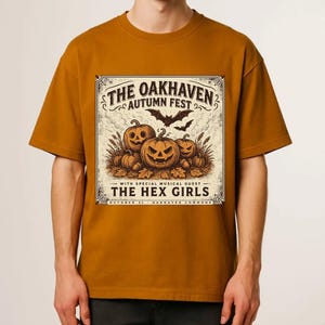 May include: A mustard yellow t-shirt featuring a graphic print of three jack-o'-lanterns, bats, and text that reads: THE OAKHAVEN AUTUMN FEST, WITH SPECIAL MUSICAL GUEST THE HEX GIRLS, OCTOBER 31, OAKHAVEN COMMONS. The design is in black and cream.
